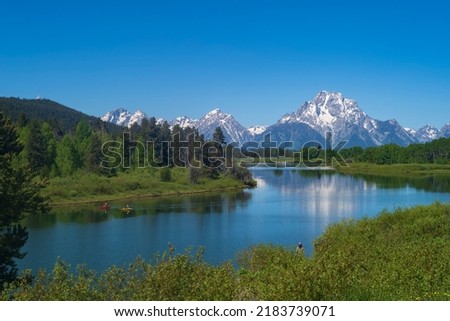 The mountain range of Grand Teton National Park is photographed during a morning with the peaks' reflections in Snake River on a cloudless summer day. 