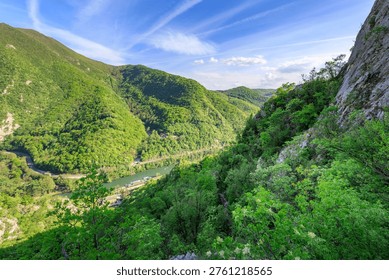 Mountain range with a river running through it. The sky is clear and blue. The lush green trees and the river create a serene and peaceful atmosphere