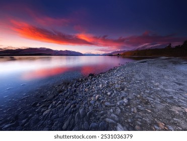 A mountain range with snow-capped peaks reflects on a calm lake with a vibrant, pink and orange sky at sunset. The lake's edge is lined with a rocky shore. - Powered by Shutterstock