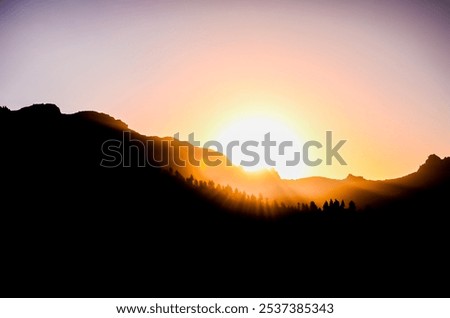 A mountain range with a sun setting behind it. The sun is low in the sky and casting a warm glow over the mountains. The scene is peaceful and serene, with the sun's rays illuminating the landscape