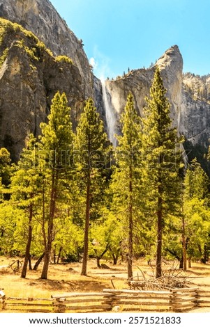 A mountain range with a waterfall in the foreground. The trees are green and the sky is blue. The scene is peaceful and serene