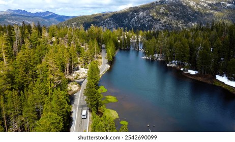 Mountain Road by Scenic Lake and Forest. Aerial view of a winding mountain road alongside a tranquil lake surrounded by dense pine forest and distant snowy peaks under a cloudy sky. - Powered by Shutterstock