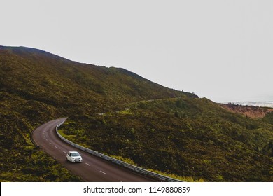 Mountain Road That Leads To Lake Of Fire In Azores. A Beautiful Green Landscape With A Single White Car On A Sinuous Road.