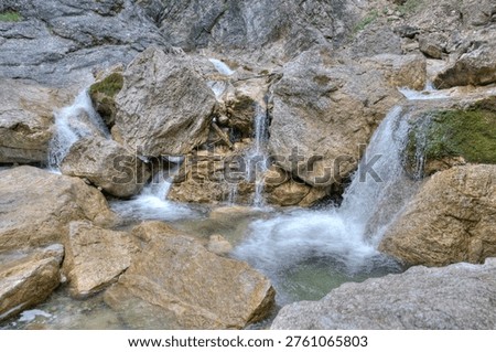 Mountain stream waterfall cascading over large boulders and rocks in alpine gorge with HDR tone mapping. Crystal clear white water rushing between mossy stones and granite rocks in natural canyon landscape. Multiple water streams converging into a small pool below dramatic rocky cliff face. Fresh mountain water with silky smooth long exposure effect on rocks covered in green moss. Perfect for nature landscape photography alpine wilderness water element themed content travel adventure outdoor recreation and environmental conservation projects.