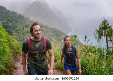 Mountain trek hikers couple walking in rain trekking on trail trek with backpacks healthy active lifestyle. Hiker man happy on hike in nature landscape with Asian girl, Na Pali Coast, Kauai, Hawaii.