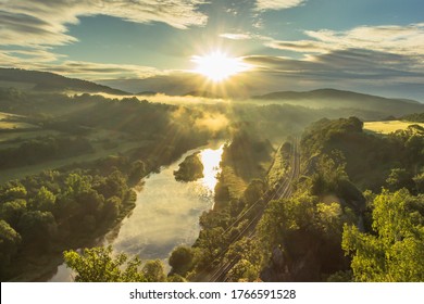 Mountain Valley During Sunrise. Natural Summer Scenic Landscape With Sun Rays. Beautiful View Of Green Valley And Railway.Sunny Day By Water. Foggy Morning Landscape Calm Atmosphere