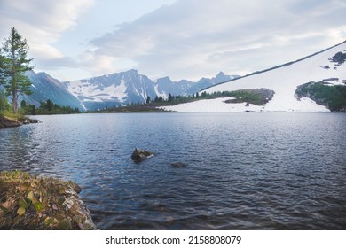 Mountain valley with snow, forest and lake at foot. Rocks are reflected in river. Melting glacier on slopes of ridge. Dawn clouds foreshadow precipitation. - Shutterstock ID 2158808079
