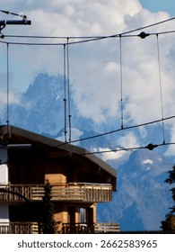 Mountain view with a large mountain range in the background. In the foreground are the roof of a wooden chalet and tangled electric wires.
