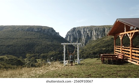 mountain view with a peaceful wooden swing framed by a white gazebo overlooking lush green hills and cliffs next to a rustic cabin in a picturesque countryside. A young girl swings at high altitude  - Powered by Shutterstock