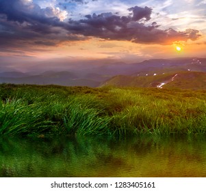 Mountain view at sunset or sunrise. Panorama of green grassy valley reflected in clear lake water, foggy mountains range with snow patches, dramatic cloudy sky and setting on horizon bright sun. - Powered by Shutterstock