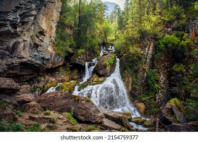 Mountain waterfall in a rocky gorge overgrown with green forest. Stream of icy water falls on mossy stones.