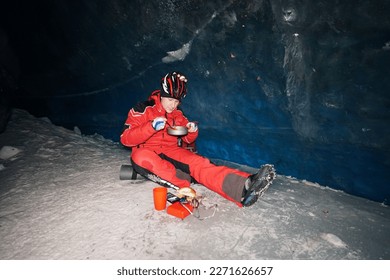 A mountaineer caver eats in a deep ice cave. Pours hot tea from a thermos, steam is coming. Eats from a frying pan, enjoys the food. Behind is a huge wall of ice with a dark blue shade. In mountains