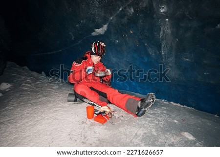 A mountaineer caver eats in a deep ice cave. Pours hot tea from a thermos, steam is coming. Eats from a frying pan, enjoys the food. Behind is a huge wall of ice with a dark blue shade. In mountains