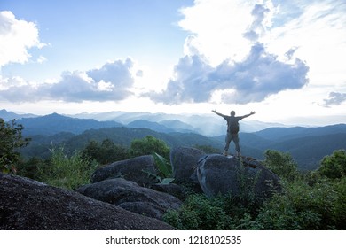 Mountaineer celebrating reaching the summit of a high rocky peak standing with his arms raised looking out over a vista of mountain ranges - Powered by Shutterstock