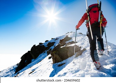 Mountaineer walking up along a snowy ridge with the skis in the backpack. In background a shiny bright sun. Concepts: adventure, courage, determination, self-realization, dangerous, extreme sport.