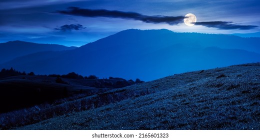 Mountainous Countryside Landscape At Night. Grassy Meadows And Trees On Hills Rolling In To The Distant Ridge In Full Moon Light