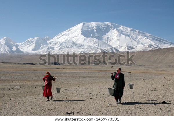 Mountainous\
landscape. Two Kyrgyz women with buckets of water on a yoke on the\
background of Muztagh Ata mountain (7546 m). Pamir mountains,\
outskirts of Kashgar, Xinjiang, China,\
Asia.