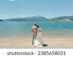 Mountains and blue water background to married couple standing on beach with bride