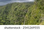 The mountains are covered with green tropical vegetation. The stream of the waterfall flows down the slope of the cliff. Clouds in the sky. Mauritius.Black River Gorges National Park.Gorges viewpoint 