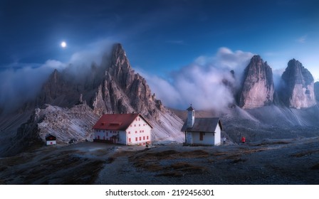 Mountains in fog with beautiful house and church at night in autumn. Landscape with high rocks, blue sky with moon. Rocky mountain peaks in clouds. Tre Cime in Dolomites, Italy. Alps at sunset in fall - Powered by Shutterstock