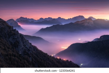 Mountains in fog at beautiful night in autumn in Dolomites, Italy. Landscape with alpine mountain valley, low clouds, forest, purple sky with stars, city illumination at sunset. Aerial. Passo Giau - Shutterstock ID 1233564241
