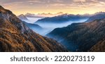 Mountains in low clouds at sunset in autumn in Dolomites, Italy. Landscape with alpine mountain hills in fog, orange trees and grass in fall, colorful sky with golden sunbeams. Aerial view. Panorama