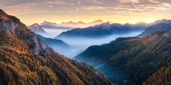 Mountains In Low Clouds At Sunset In Autumn In Dolomites, Italy. Landscape With Alpine Mountain Hills In Fog, Orange Trees And Grass In Fall, Colorful Sky With Golden Sunbeams. Aerial View. Panorama