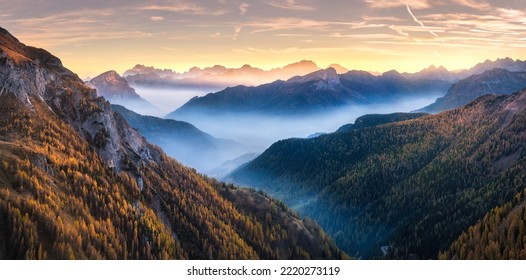 Mountains in low clouds at sunset in autumn in Dolomites, Italy. Landscape with alpine mountain hills in fog, orange trees and grass in fall, colorful sky with golden sunbeams. Aerial view. Panorama - Powered by Shutterstock