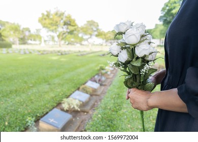 Mourning young woman holding white flowers at her family grave in beautiful green cemetery.