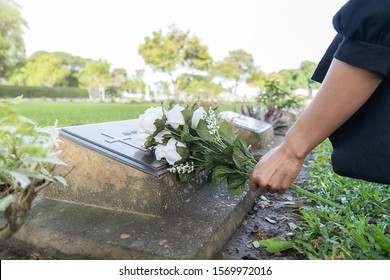 Mourning young woman laying white flowers on her family grave in beautiful green cemetery.