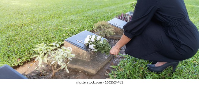 Mourning young woman laying white flowers on her family grave in beautiful green cemetery.