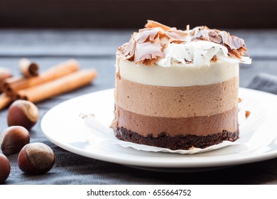 Mousse Cake Three Chocolate, Whole Hazelnut And Cinnamon Sticks On A White Plate. Dark Wooden Background.