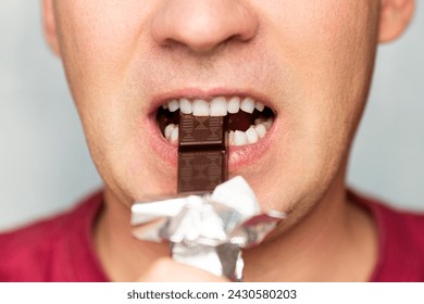 Mouth of man eating chocolate against blue background. A man's mouth in close-up with white teeth biting dark chocolate. - Powered by Shutterstock