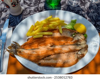 mouthwatering plate of grilled fish, served with a side of crispy French fries, fresh tomato slices, and a lemon wedge. Golden brown crust and is garnished with onion rings. Traditional restaurant - Powered by Shutterstock