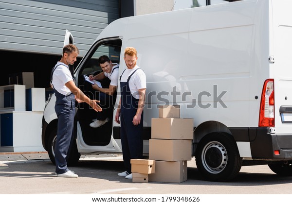Movers in overalls standing near cardboard boxes\
and truck on urban\
street