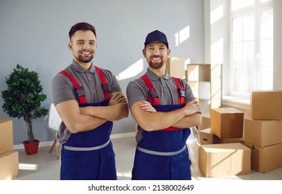 Movers team. Two young cheerful employees of moving service in overalls are smiling in front of camera. Portrait of men posing with crossed arms standing between cardboard boxes in apartment.