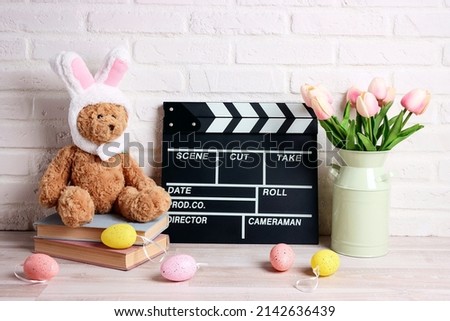 Movie clapper board with Easter decorations against a white brick wall. Cinema for the spring holidays. Funny toy bear with rabbit ears, tulips and colored eggs.