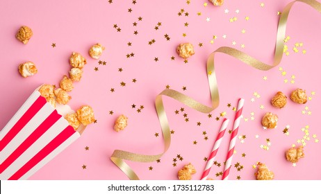 Movie theater party or birthday in cinema concept. Flat lay composition with red and white striped box with popcorn and drink straw, golden party streamers and confetti stars on pink table. - Powered by Shutterstock