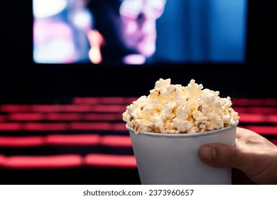 Movies and popcorn. Man holding pop corn box at cinema. Action, thriller or scifi entertainment on screen. Red seats in dark theater. Salty snack in bucket. Spectator pov. Film premiere.