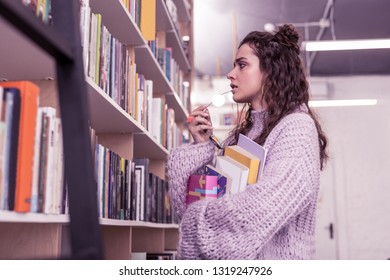 Moving All Around. Serious Young Woman With Wavy Hair Unfolding Her Clear Glasses For Better View On Book Titles