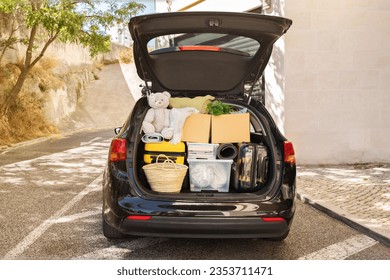Moving boxes and suitcases, various belongings, plants in open trunk of black car outdoor on the street next to building, no people, copy space. Relocation, moving, real estate, new beginning concept