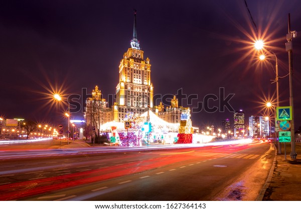 Moving\
car with blur light through city at night near Moscow river\
embankment and illuminated Radisson or Ukraine hotel. Moscow,\
Russia. Happy new year 2020 decoration in\
Russian.