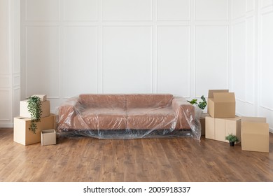 Moving Day Concept. Couch Under Polyethylene Cover, Cardboard Carton Boxes Stack With Household Belongings In Modern House Living Room, Packed Containers On Floor In New Home. Relocation, Renovation