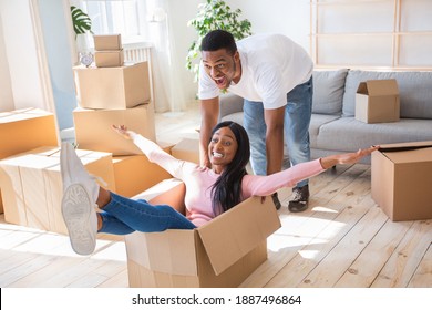 Moving Day Fun. Millennial Newlywed Black Couple Being Silly And Playful In Their New House, Riding Carton Box. Happy Young Woman And Her Husband Playing Game During Relocation