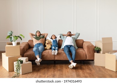 Moving Day. Lovely Arab Family Relaxing On The Couch In New Home With Cardboard Boxes Around. Happy Man, Woman In Headscarf And Little Girl Lying On Sofa And Looking Up, Free Copy Space