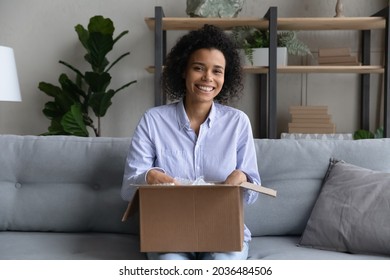 Moving day. Portrait of excited young biracial woman realty buyer renter sit on sofa hold big box pack belongings for relocation to new home. Happy black lady look at camera get order from online shop