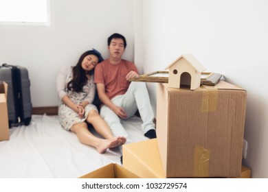 Moving To New Blank House. Tired Young Asian Family Couple Sleeping Together Near Many Cardboard Boxes And Luggage. Selective Focus At Wooden House Model And Photo Frame. Start New Life.