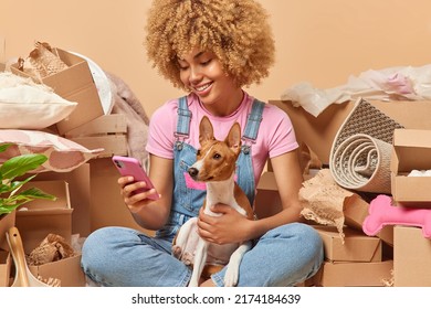 Moving to new home concept. Positive curly haired woman checks newsfeed or makes orders online on smartphone poses with dog in messy room surrounded by carton boxes relocates to new place of living