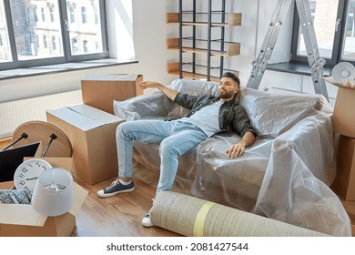 Moving, People And Real Estate Concept - Tired Man With Boxes Resting On Sofa Covered With Plastic Sheeting At New Home