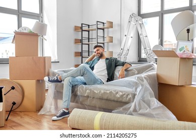 Moving, People And Real Estate Concept - Happy Smiling Man With Boxes Resting On Sofa Covered With Plastic Sheeting At New Home